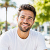 Man with a beard and short hair wearing a white t-shirt, smiling outdoors with greenery in the background.